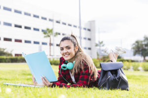 portrait-smiling-female-university-student-lying-green-grass-holding-book-hand (1)